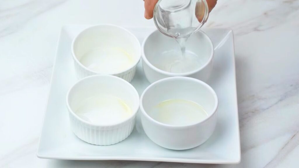 Hot water being poured into small white ramekins on a plate, preparing the cups for making air fryer poached eggs.