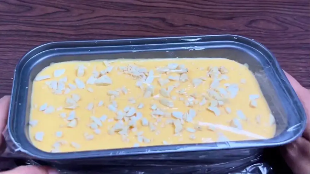Loaf pan filled with 3 ingredient mango ice cream mixture, topped with chopped nuts and covered with plastic wrap before freezing for storage.
