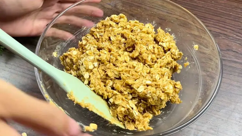 Peanut butter oatmeal cookie dough mixed in a glass bowl, with rolled oats evenly coated and the dough holding together in thick, textured clumps.