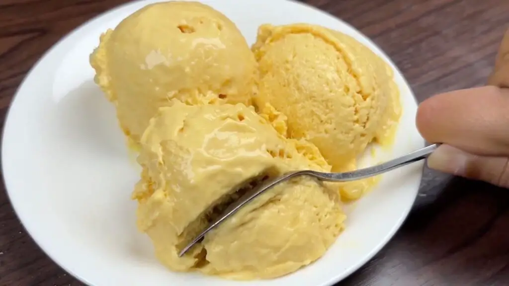 Scoops of creamy homemade mango ice cream in a white bowl, with a spoon lifting a soft, smooth bite to show the rich texture of this easy mango ice cream recipe.