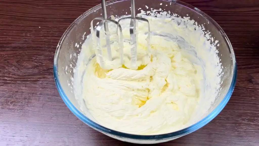 Thick whipped cream being mixed with an electric hand mixer in a glass bowl during the process of making homemade coconut ice cream.