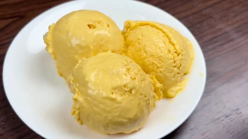 Three scoops of creamy mango ice cream served on a white plate, showing the smooth texture and rich golden mango color of this homemade mango ice cream.
