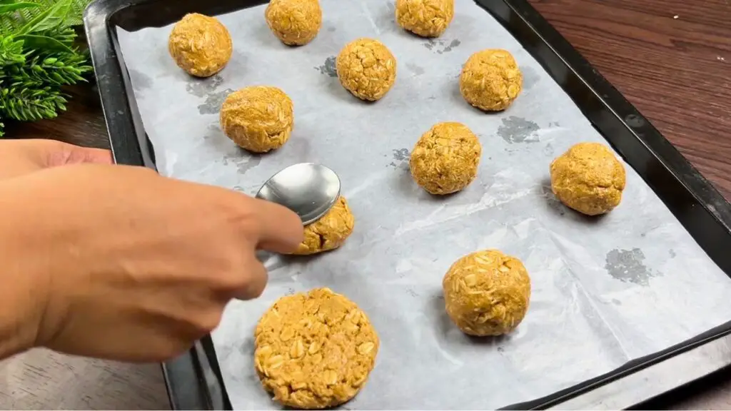Using the back of a spoon to flatten peanut butter oatmeal cookie dough balls on a parchment-lined baking tray, shaping them into evenly sized cookies before baking.