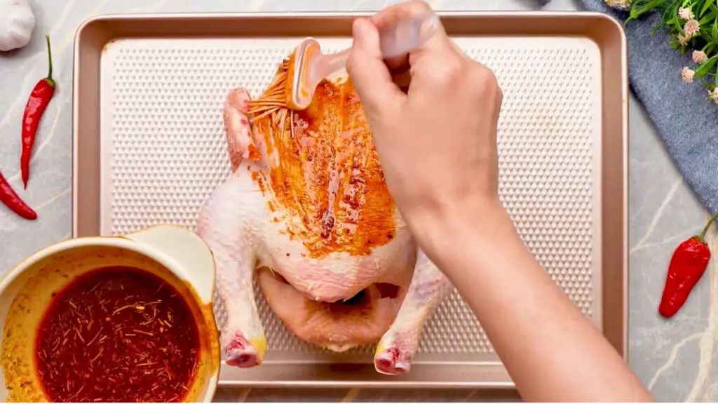 Whole raw chicken being brushed with a seasoned marinade on a baking tray, preparing it for air fryer whole chicken cooking.