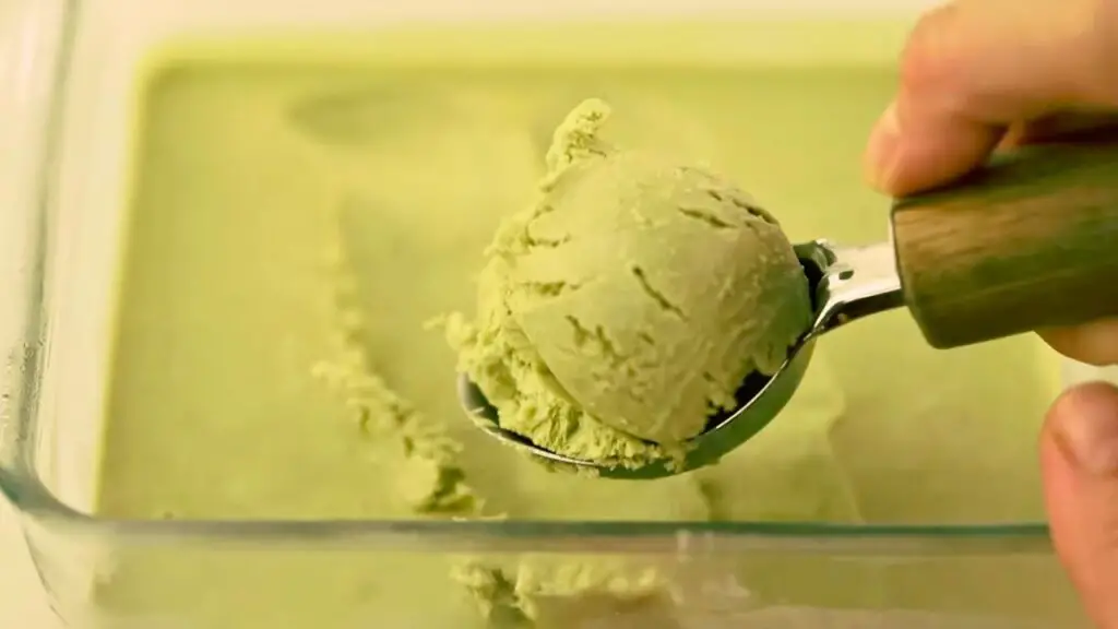 A scoop of homemade matcha green tea ice cream being lifted from a freezer-safe container, showing its smooth, creamy texture.