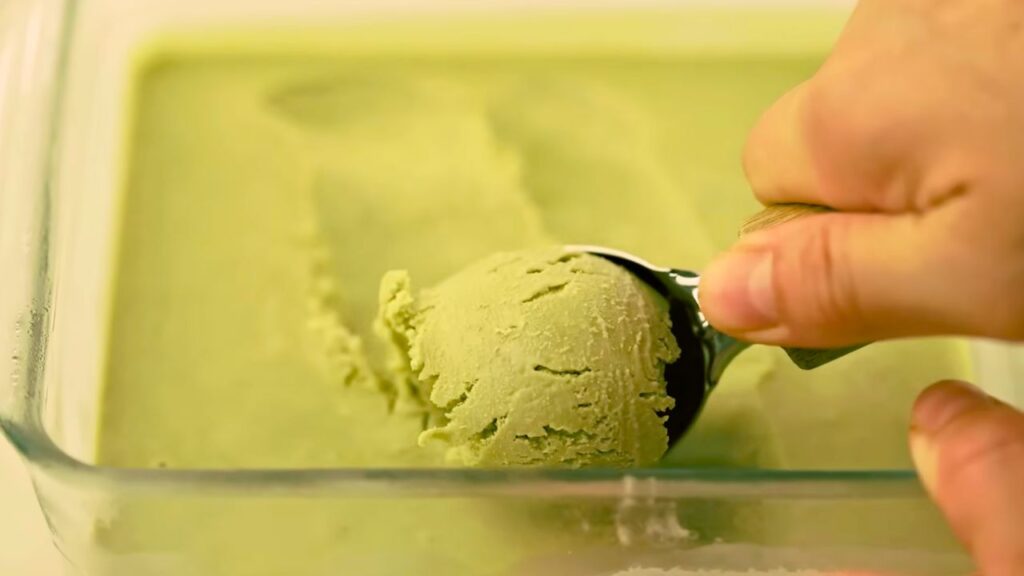 A scoop of homemade matcha ice cream being served from a glass freezer-safe container, showing its smooth, creamy texture.