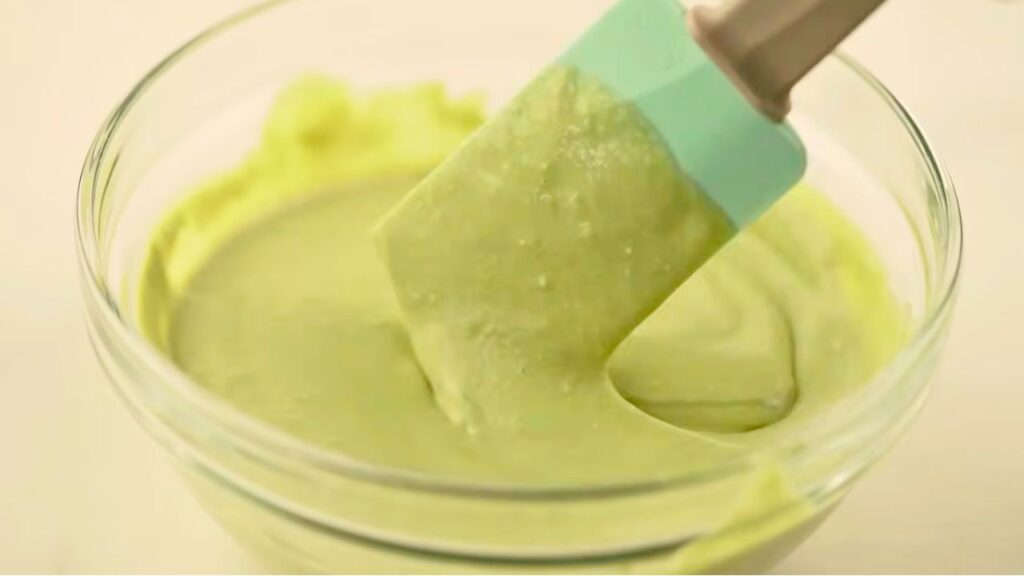 A spatula mixing smooth pale green matcha ice cream mixture in a glass bowl until the matcha is fully blended.