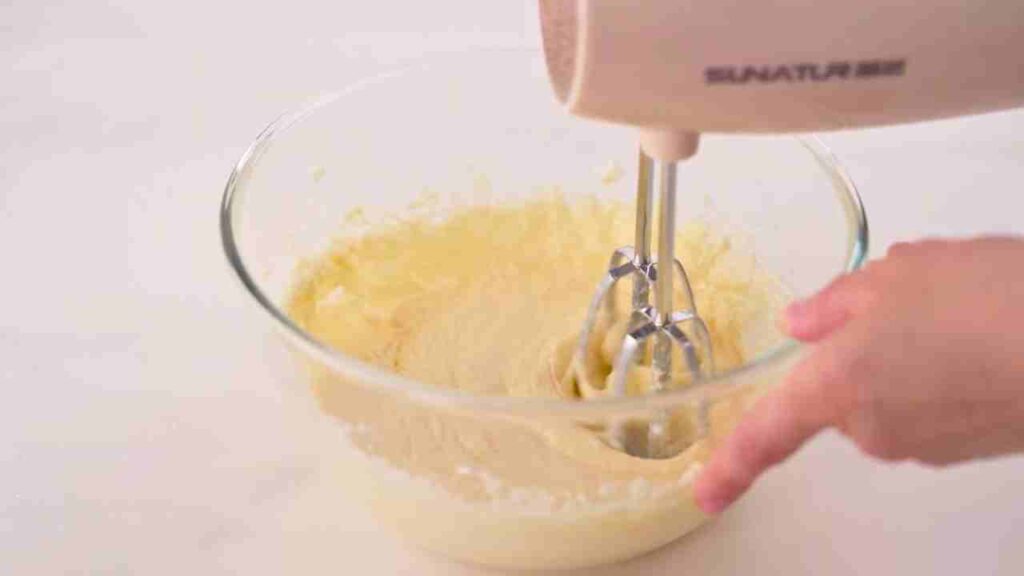 Apple waffle recipe showing eggs being mixed into creamed butter and sugar with an electric hand mixer in a glass bowl to create a smooth waffle batter.