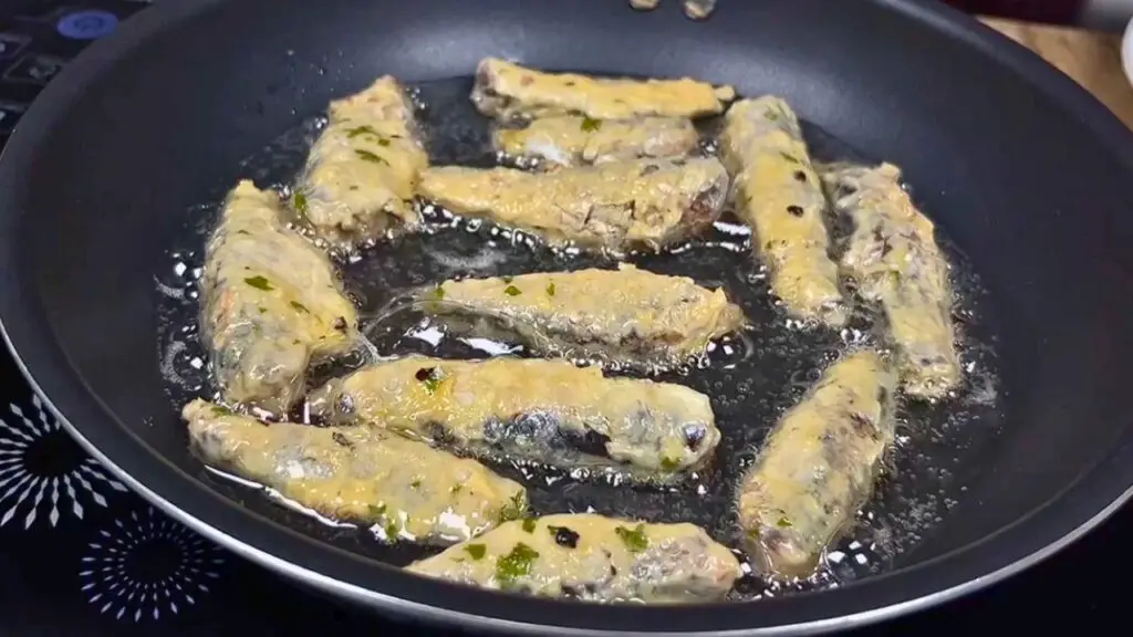 Canned sardines fried in a skillet with a light golden coating, cooking in hot oil until crispy on the outside.