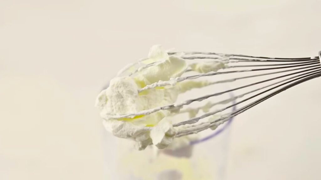 Close-up of whipped cream on a whisk showing soft peaks, ready for making homemade matcha ice cream.