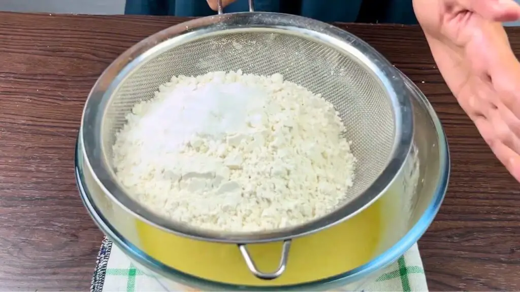 Dry ingredients being sifted through a fine-mesh sieve into wet batter while making an easy homemade lemon loaf cake.