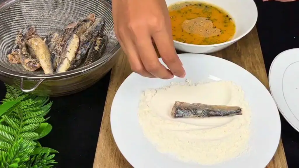 Fried sardines recipe showing a sardine being lightly coated in flour before dipping into the egg mixture for a crispy coating.