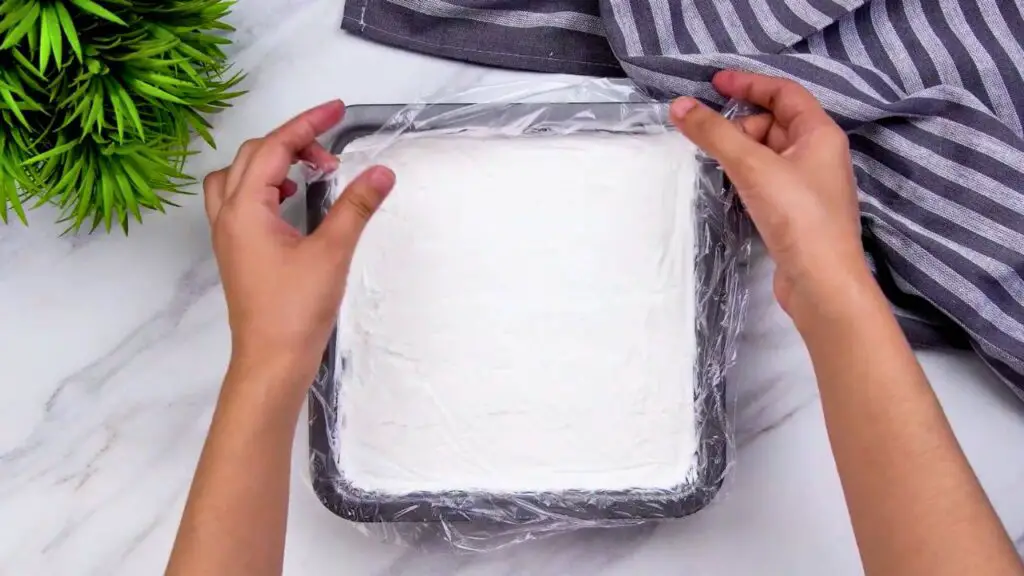 Hands removing the plastic wrap from homemade Greek yogurt ice cream after freezing in a square freezer-safe pan.