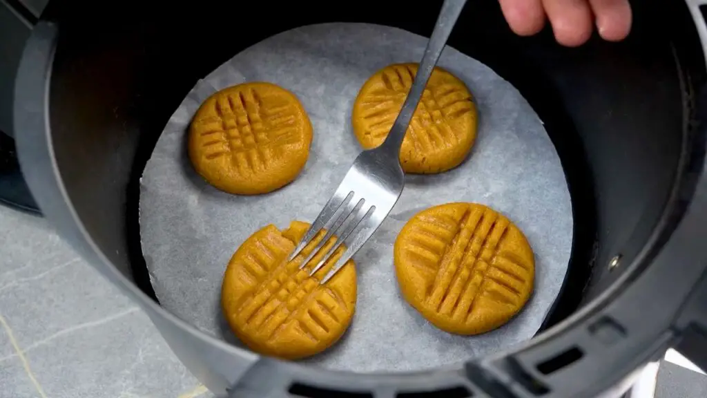 Peanut butter cookie dough in an air fryer basket with a fork pressing the classic criss-cross pattern on top.
