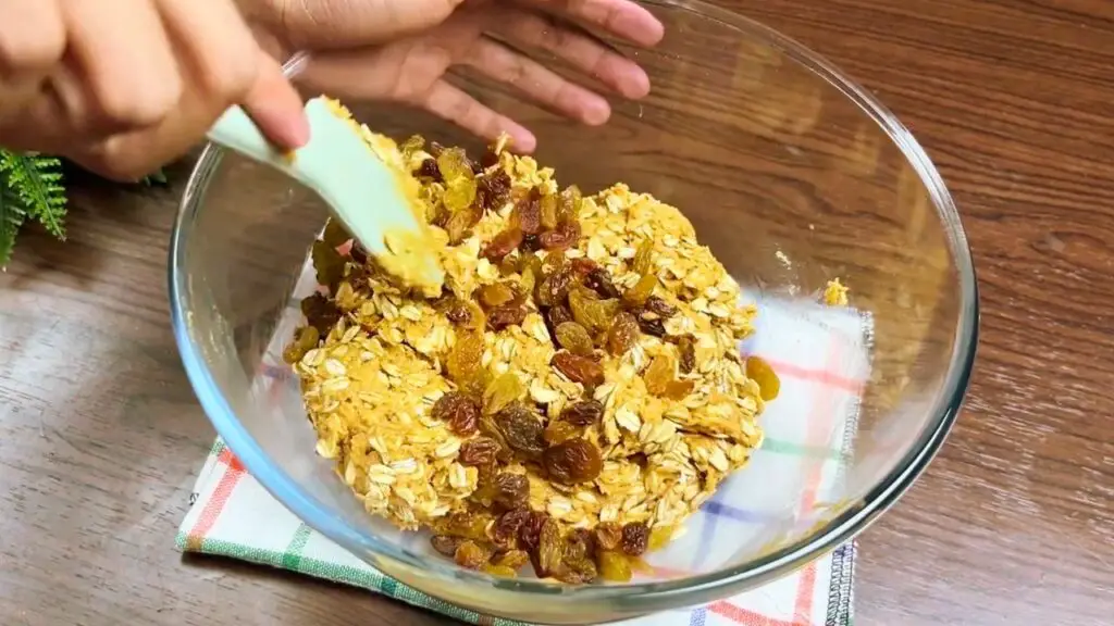 Peanut butter oatmeal cookies with raisins being folded into the oat cookie dough in a glass mixing bowl.