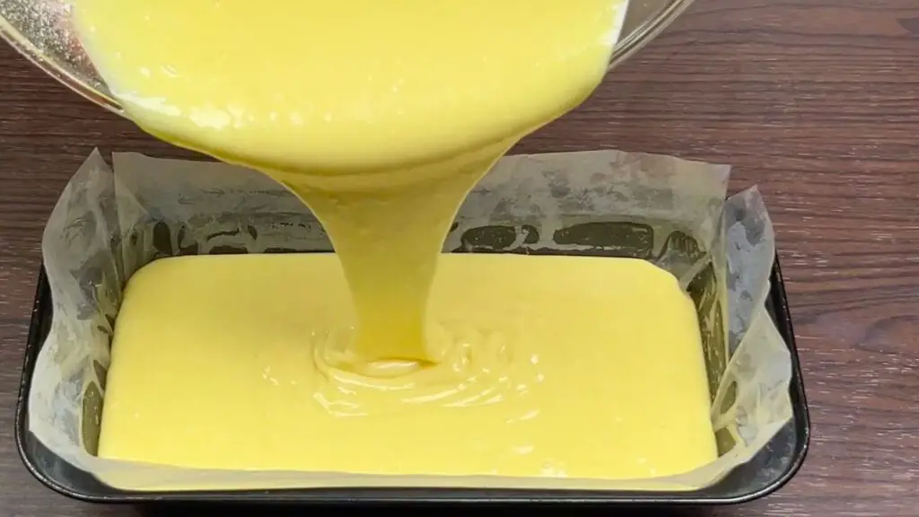 Pouring sweetened condensed milk cake recipe batter into a parchment-lined loaf pan before baking.