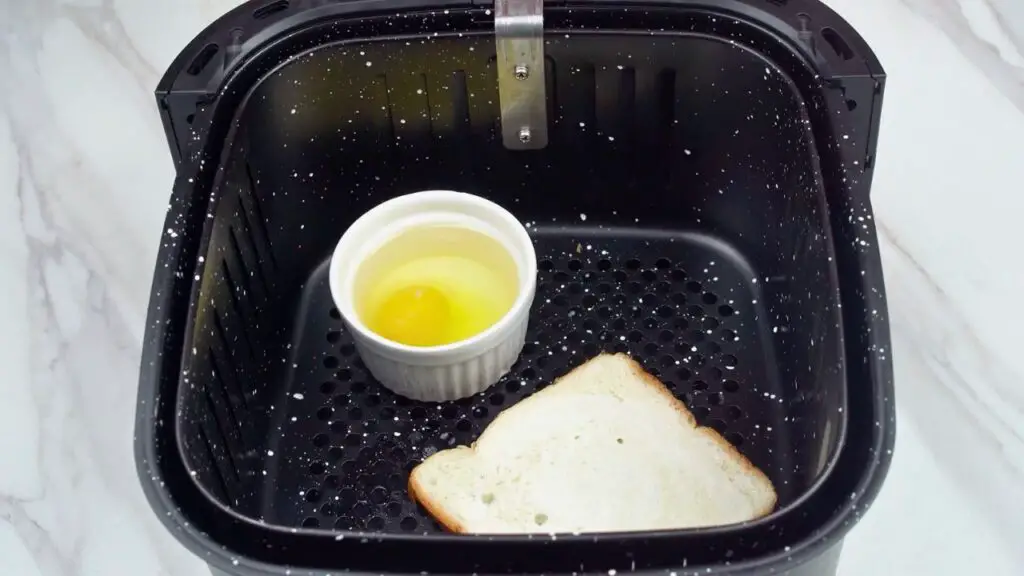 Ramekin with a raw egg and slice of bread in an air fryer basket before cooking poached eggs in air fryer.