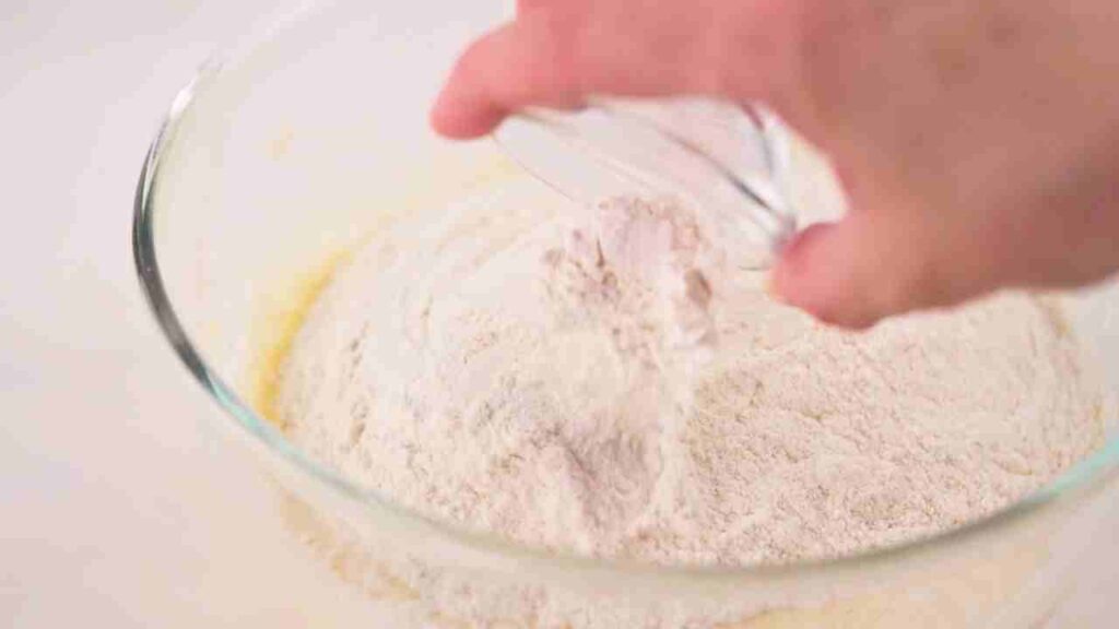Waffle maker recipe showing all-purpose flour, baking powder, and salt being added to the wet ingredients in a glass mixing bowl before gently mixing into a smooth waffle batter.