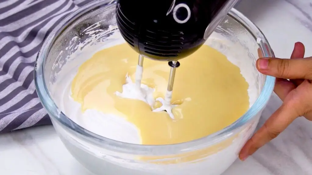 Yogurt ice cream mixture with whipped cream and sweetened condensed milk being blended together using a hand mixer.