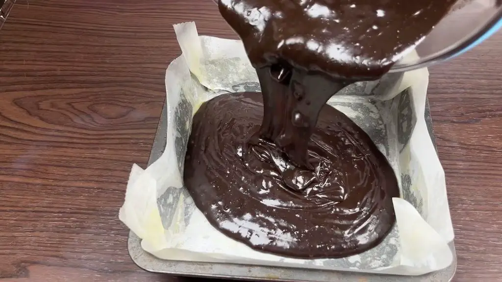 Fudgy homemade brownies batter being poured into a parchment-lined baking pan before baking.