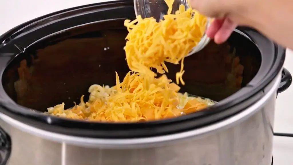 Shredded sharp cheddar and cubed cheese being added to slow cooker mac n cheese for a rich and creamy cheese sauce.