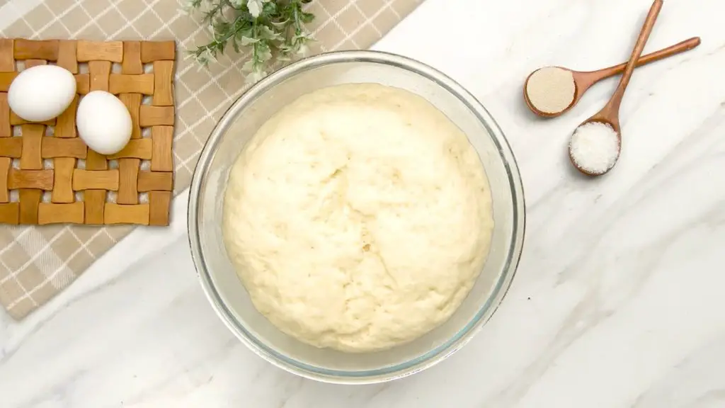Brioche bun dough doubled in size in a glass mixing bowl after rising.