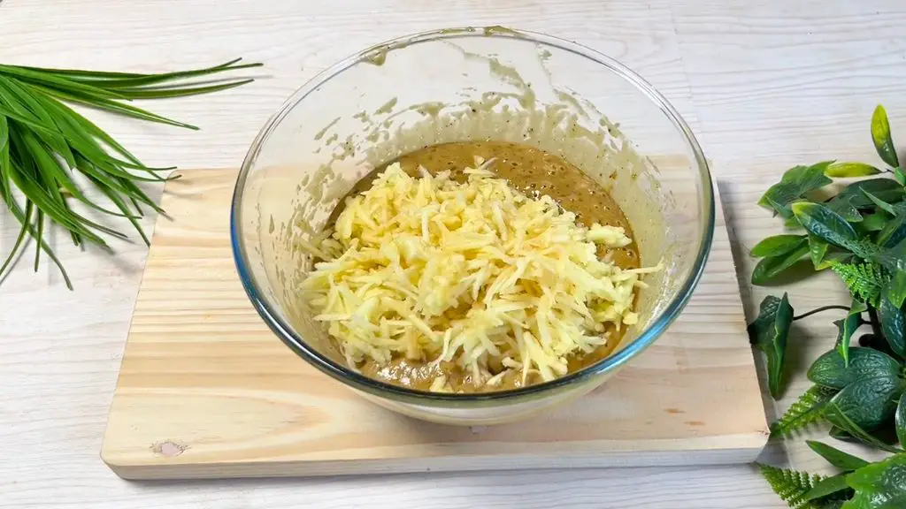Grated apples added to a glass bowl of batter for a toffee apple cake recipe, ready to be mixed with chopped walnuts.