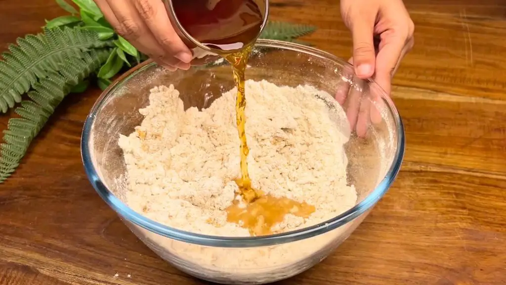 Leftover black tea from soaked raisins and sultanas being poured into a glass bowl of flour mixture for a traditional fruit tea loaf recipe.