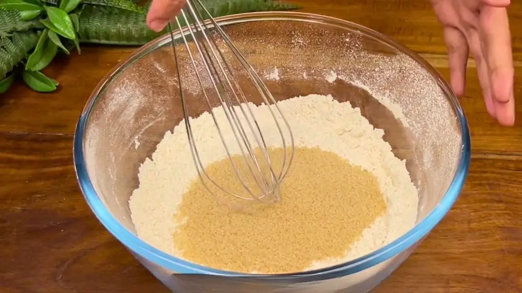 Light brown sugar being added to a glass bowl of flour and spices, ready to be mixed with a whisk for a traditional fruit cake recipe.