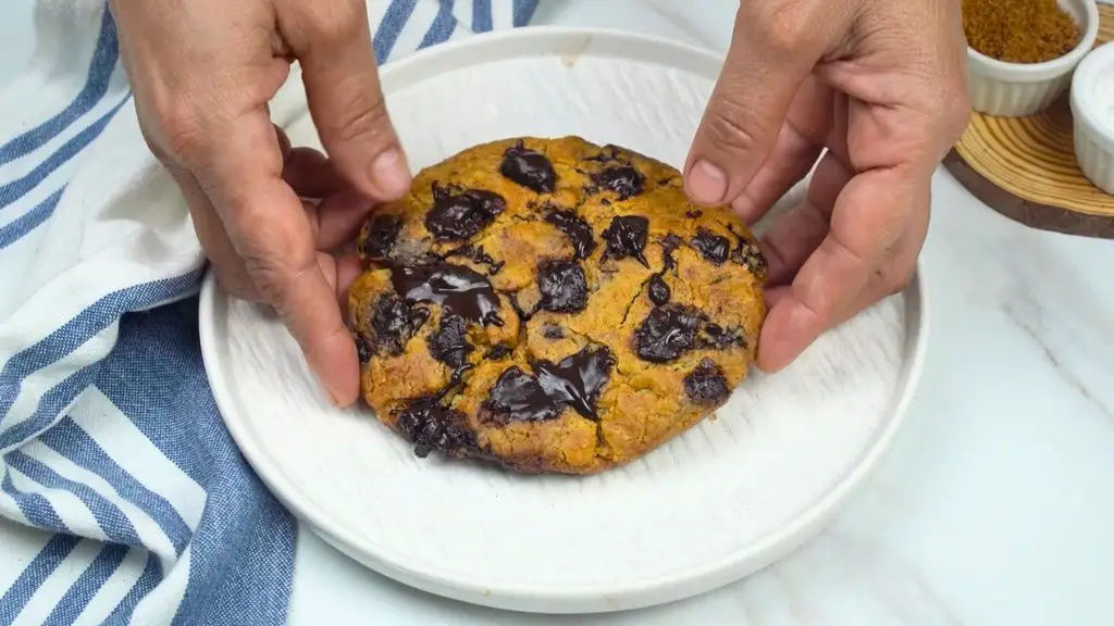 Single serve air fryer chocolate chip cookie with a golden brown crust and melted chocolate chunks, being placed on a white plate.
